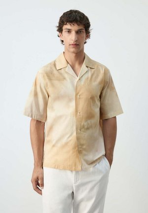 Junger Mann mit kurzen lockigen Haaren trägt ein beige-hellbraunes kurzärmliges Hemd mit Knöpfen und weiße Hosen vor einem einfarbigen Hintergrund.