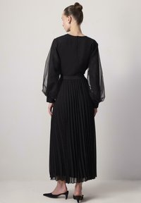 Zwarte lange jurk met doorschijnende pofmouwen, geplooide rok en een nauwsluitende taille. Gesloten rug met een rits en een lage halslijn.