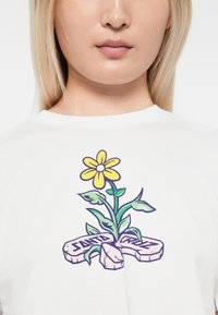 Hvit t-skjorte med et fargerikt blomsterdesign med gule kronblader og grønne blader, akkompagnert av steiner merket "SANTA CRUZ."