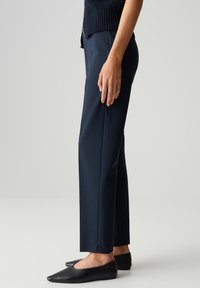 Pantalon marine sur mesure avec une coupe droite, fabriqué dans un tissu texturé. Caractéristiques d'une fente latérale au bas et associé à des chaussures plates pointues.