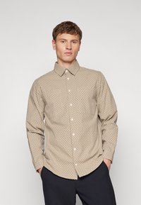 Beige gombos ing geometrikus mintával, hosszú ujjakkal és hegyes gallérral. Az anyag sima és könnyűnek tűnik.