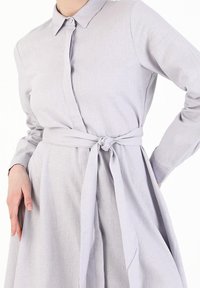 Robe gris clair avec un col, un bouton devant et des manches longues. Comprend une taille cintrée et une jupe fluide. Tissu lisse et texturé.