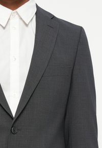 Giacca di suit grigia su misura con una superficie liscia, chiusura a un bottone e revers a punta, abbinata a una camicia bianca con bottoni.