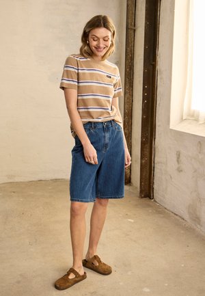 Kvinde står indendørs iført en beige t-shirt med striber, knælange blå denimshorts og brune slip-on sko, smilende og kigger ned.