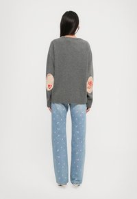 Gray sweater with a relaxed fit, ribbed hem, and patch elbow details featuring red designs. Paired with light blue jeans adorned with floral patterns.