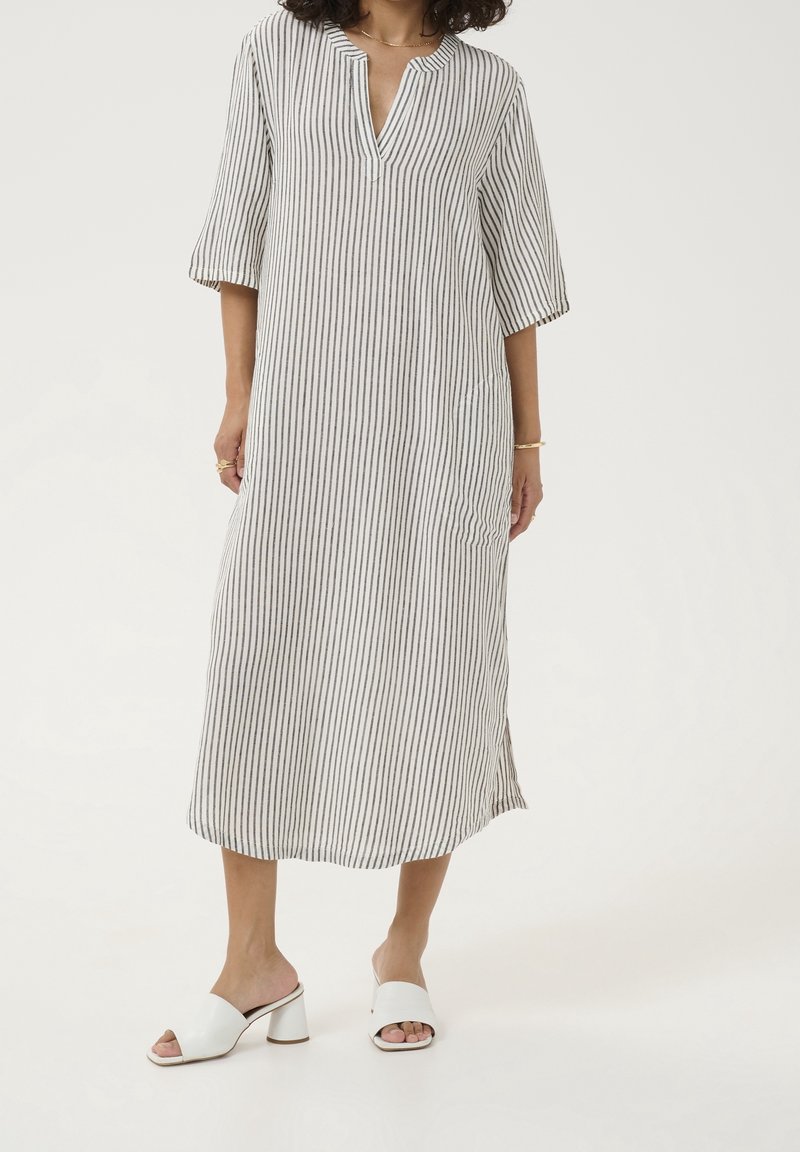 Femme portant une robe midi blanche avec fines rayures noires verticales, manches trois quarts, col en V et chaussures blanches à talons blocs ouvertes à l'avant.
