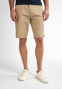 Pantaloni scurți bej din bumbac, cu un design plat în față, cu tăietură până la genunchi și buzunare laterale subtile, asortați cu adidași albi.