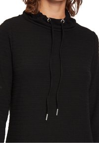 Svart topp med cowl-neck har en texturerad ribbat design med dragsko och silverfärgade spetsar. Tyget verkar mjukt och något stretchigt.