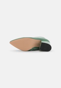 Zapato de tacón alto verde con acabado brillante, punta afilada, suela beige y una sección de tracción negra en el talón.
