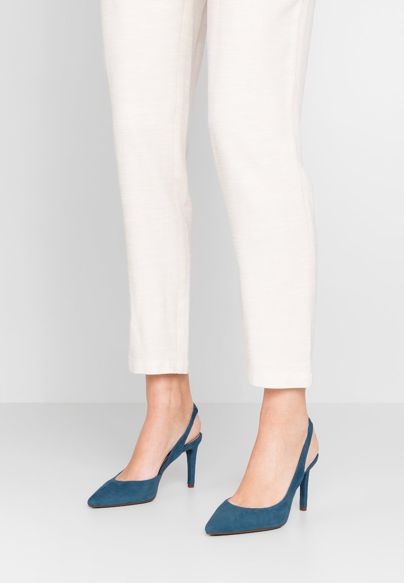 Chaussures slingback à talons hauts en daim bleu sarcelle, avec un bout pointu, un talon fin et un dos ouvert, assorties à un pantalon fuselé de couleur claire.