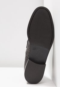 Semelle d'une chaussure habillée en cuir noir avec une surface lisse légèrement texturée, reposant verticalement contre un mur blanc d'angle.