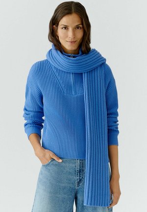 Blaues geripptes Strickpullover mit hochgeschlossenem Kragen und passendem Schal. Der Schal hat ein strukturiertes Design und hängt herunter, was den Pullover ergänzt.