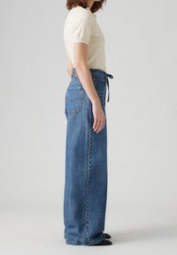 Femme portant un haut crème à manches courtes rentré dans un jean bleu taille haute à jambes larges avec une ceinture à cordon noire et des chaussures noires.