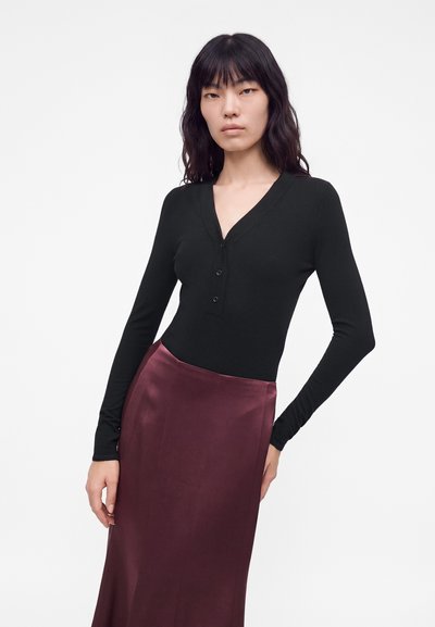 Femme aux longs cheveux noirs portant un haut noir à manches longues avec des boutons et une jupe taille haute en satin violet foncé sur fond blanc.