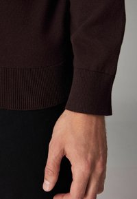 Tejido de suéter marrón oscuro con puños y dobladillo acanalados. Primer plano de una mano apoyada sobre la tela, mostrando una textura de piel suave.
