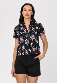 Blouse noire à manches courtes avec un motif floral rose, dotée d'un col et d'une fermeture à boutons, assortie de shorts noirs ajustés.