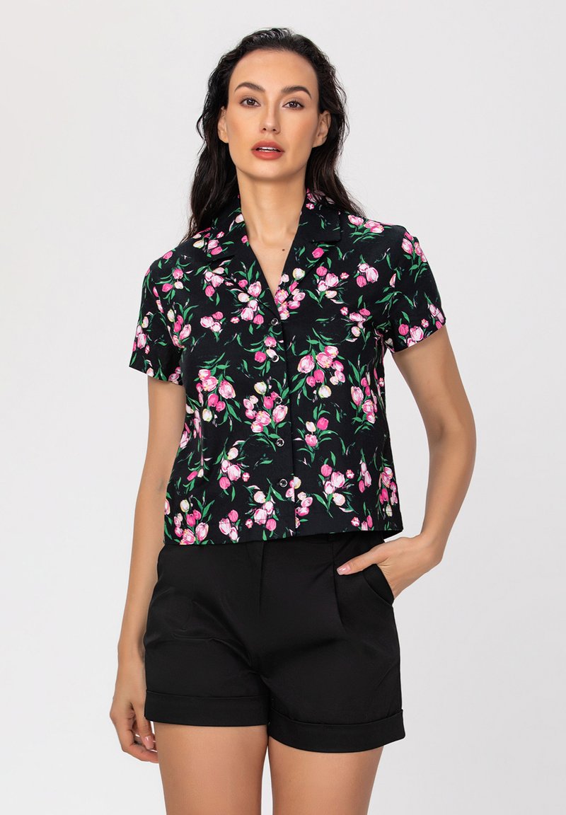 Blouse noire à manches courtes avec un motif floral rose, dotée d'un col et d'une fermeture à boutons, assortie de shorts noirs ajustés.