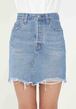 Mini-jupe en denim bleu clair avec ourlet effiloché et détails asymétriques à l'avant, portée avec un haut blanc rentré.