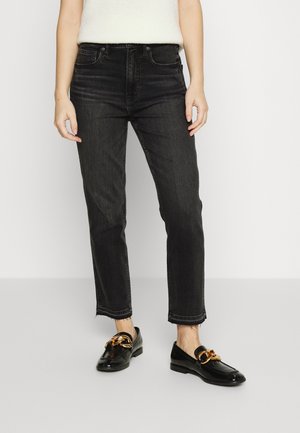 Person iført højtaljede sorte jeans med rå kanter og sorte læderloafers med stort guldkæde-detalje på hvid baggrund.