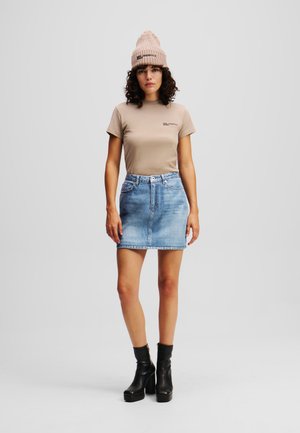 T-shirt beige con logo, mini gonna di jeans blu chiaro e stivali neri con tacco alto alla caviglia. Il modello indossa un berretto di maglia rosa chiaro.