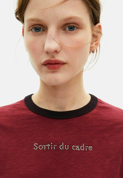 Burgunder T-Shirt mit schwarzem geripptem Rundhalsausschnitt, auf der Vorderseite mit silberner, genagelter Schrift "Sortir du cadre" und glatter Textur.