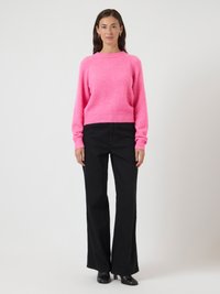 Mulher com cabelo longo e escuro, vestindo um suéter rosa brilhante, calças largas pretas, botas de salto pretas e brincos de argola dourados, de pé contra um fundo branco.