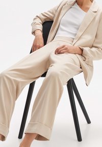 Personne assise sur une chaise noire, portant un pantalon de costume beige, un blazer assorti et un haut sans manches blanc, dans une pose détendue.