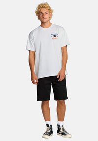 Blonde man met krullend haar, gekleed in een witte Billabong t-shirt, zwarte shorts, witte sokken en zwarte hoge sneakers op een effen achtergrond.