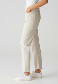 Beige rechtliggende broek met rafelige pijpen, gemaakt van denim, met zijzakken. Gepaard met witte sneakers, weergegeven in zijaanzicht.