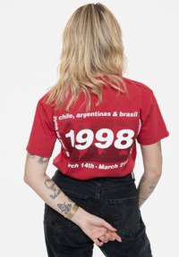 Red cotton t-shirt featuring a bold white graphic design on the back, including the year "1998" and event details, with short sleeves.