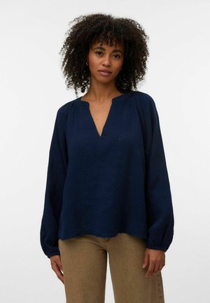 Femeie cu păr creț, care poartă o bluză largă bleumarin cu mâneci lungi și gât în V și pantaloni bej, stând în fața unui fundal alb simplu.