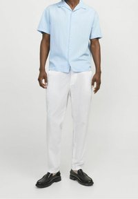 Man med ljusblå kortärmad skjorta, vita byxor och svarta loafers, stående mot en enfärgad vit bakgrund.