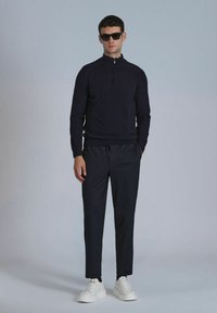 Marineblauer Reißverschluss-Pullover mit hohem Kragen, kombiniert mit dunklen Jogginghosen und weißen Sneakers. Das Model trägt schwarze Sonnenbrillen.