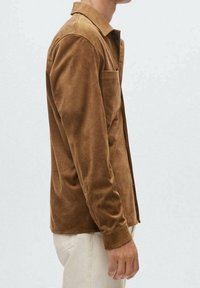 Brun ruskinds skjorte med knaplukning, lange ærmer og en enkelt brystlomme. Stofret ser blødt og struktureret ud.