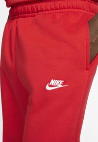 Red sweatpants made of soft fabric featuring an elastic waistband, side pockets, and a white Nike logo on the left thigh.