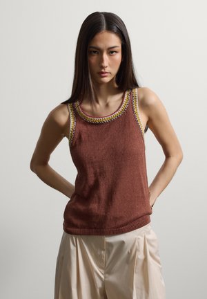 Jeune femme aux cheveux bruns et lisses portant un haut sans manches en tricot marron avec une bordure multicolore et un pantalon plissé beige, debout sur fond gris.
