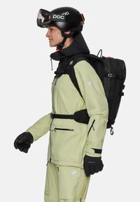 Skibekleidung mit einer hellgrünen wasserdichten Jacke mit schwarzen Akzenten, passenden Hosen, Handschuhen und einem schwarzen Rucksack. Schwarzer Helm und Skibrille.