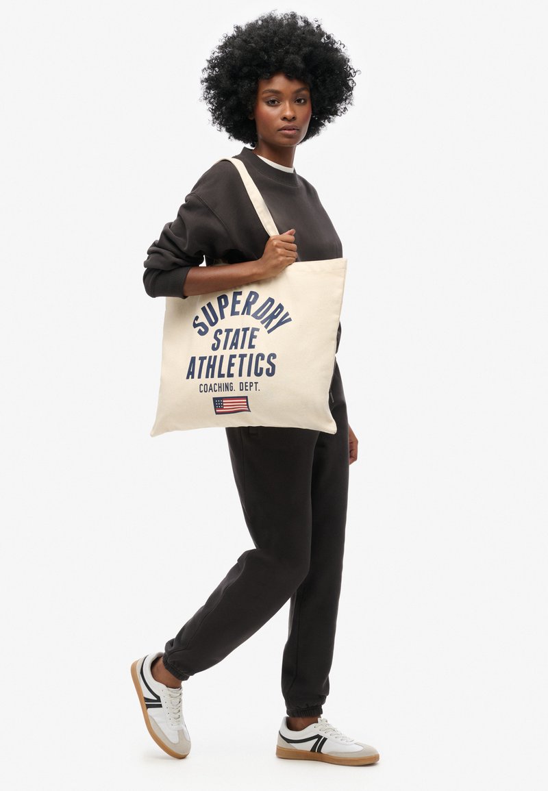 Katoenen tote bag in natuurkleur met een marineblauw gedrukt logo "SUPERDRY STATE ATHLETICS" en een kleine Amerikaanse vlag. Model in bijpassende donkere outfit.