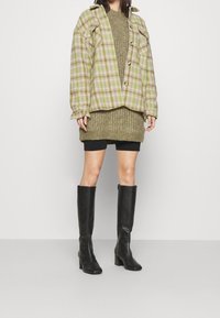 Übergroße karierte Hemdjacke in Grün und Beige, übereinander getragen mit einem kräftigen olivgrünen Pullover, kombiniert mit schwarzen Mini-Shorts und knielangen schwarzen Stiefeln.