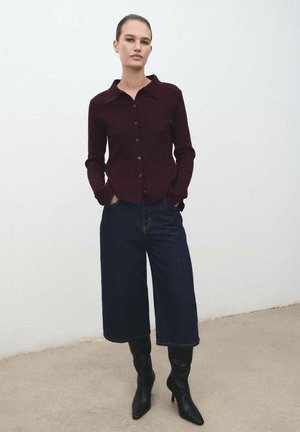 Cárdigan burdeos de botones, ligeramente ajustado, con cuello puntiagudo, combinado con jeans de pierna ancha azul oscuro y botas negras hasta la rodilla.