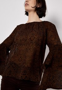 Blusa marrón con estampado, de mangas largas y acampanadas, hecha de un tejido ligero y semitransparente que presenta intrincados diseños florales.