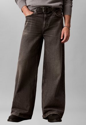 Mann trägt ausgewaschene, weite schwarze Jeans mit schwarzem Gürtel, grauen Pullover und schwarze Schuhe, steht mit einer Hand in der Tasche.