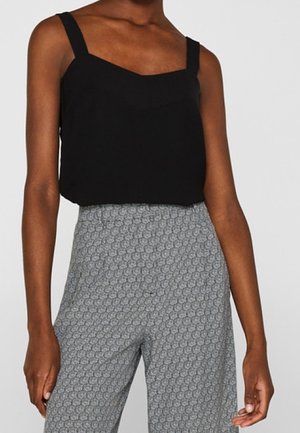 Persoon draagt een zwarte mouwloze top en hooggetailleerde grijze broek met patroon tegen een effen lichte achtergrond.