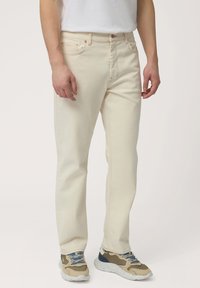Jeans de mezclilla beige con corte recto, cierre de botón y detalles de costura en naranja. Llevados con zapatillas gruesas de varios colores.