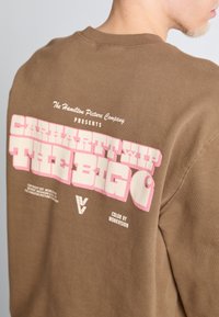 Sudadera marrón con gran texto en blanco y rosa en la parte posterior, con elementos de marca y diseño de cuello redondo. Textura de tela suave.