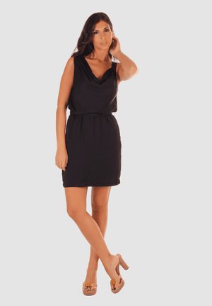 Vestido negro sin mangas con escote drapeado, cintura fruncida y dobladillo a la altura de la rodilla. La modelo lleva sandalias de tacón color beige con adornos decorativos.