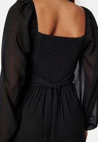 Vestito nero con schiena quadrata, dettagli arricciati sul corpetto e lunghe maniche trasparenti, con vita legata per una silhouette aderente.
