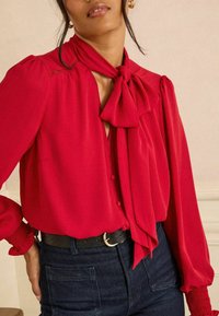 Piros blúz masnival a nyakban, puffos ujjakkal és gyűrött mandzsettával, gombos záródással. Sötét farmerrel és fekete övvel párosítva.