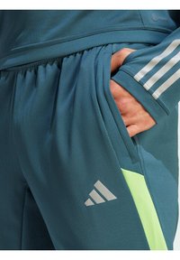 Teal sportieve joggingbroek van ademend materiaal, met een grijs Adidas-logo, een meshstructuur en een gele accentzak.