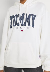 Sudadera blanca con texto "TOMMY JEANS" en navy y logo de bandera en el pecho, bolsillo frontal y capucha con cordones, llevada con pantalones oscuros.